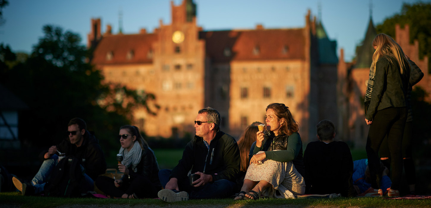 Et par mennesker sidder på græsset udenfor i aftensolen. I baggrunden ses Egeskov Slot.