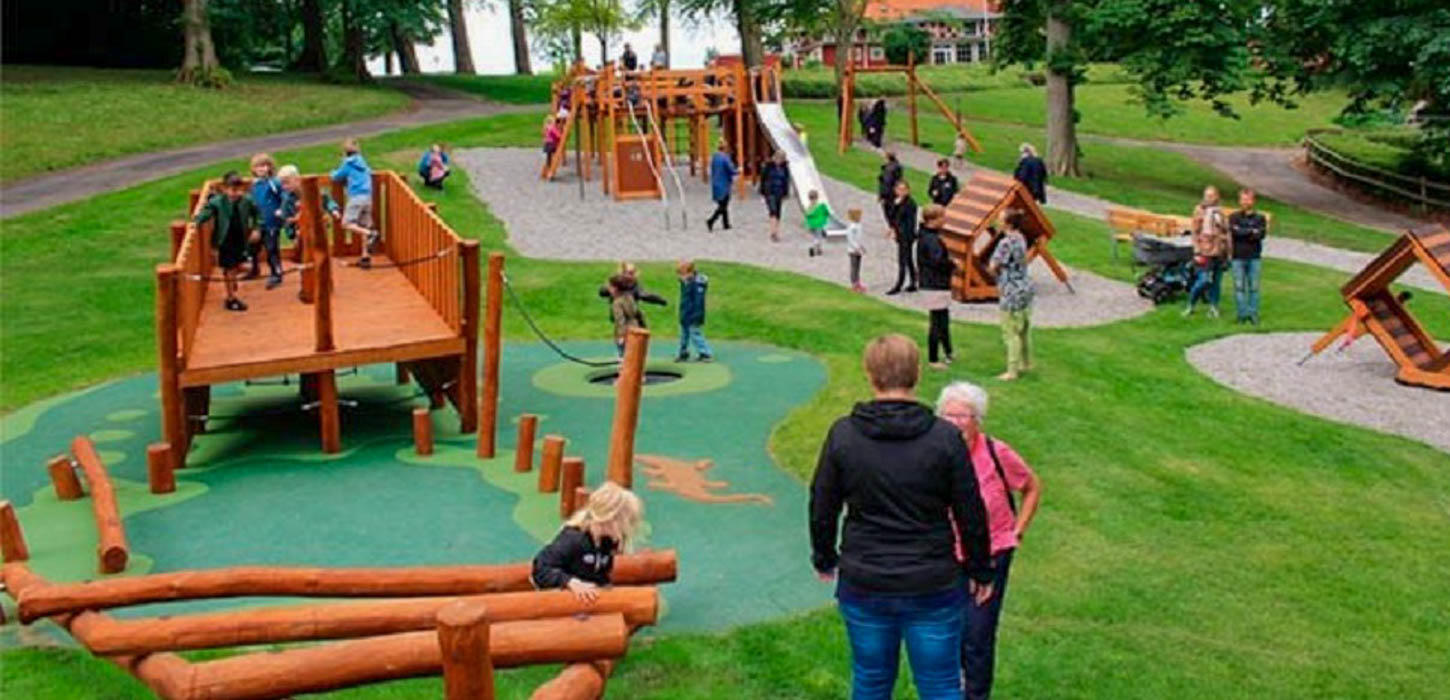 En stor græsplæne har tre steder store pletter af grus. På grus-pletterne er forskellige legepladser af træ stillet op. Rundt om og på legepladserne er der massere af børn og voksne. 