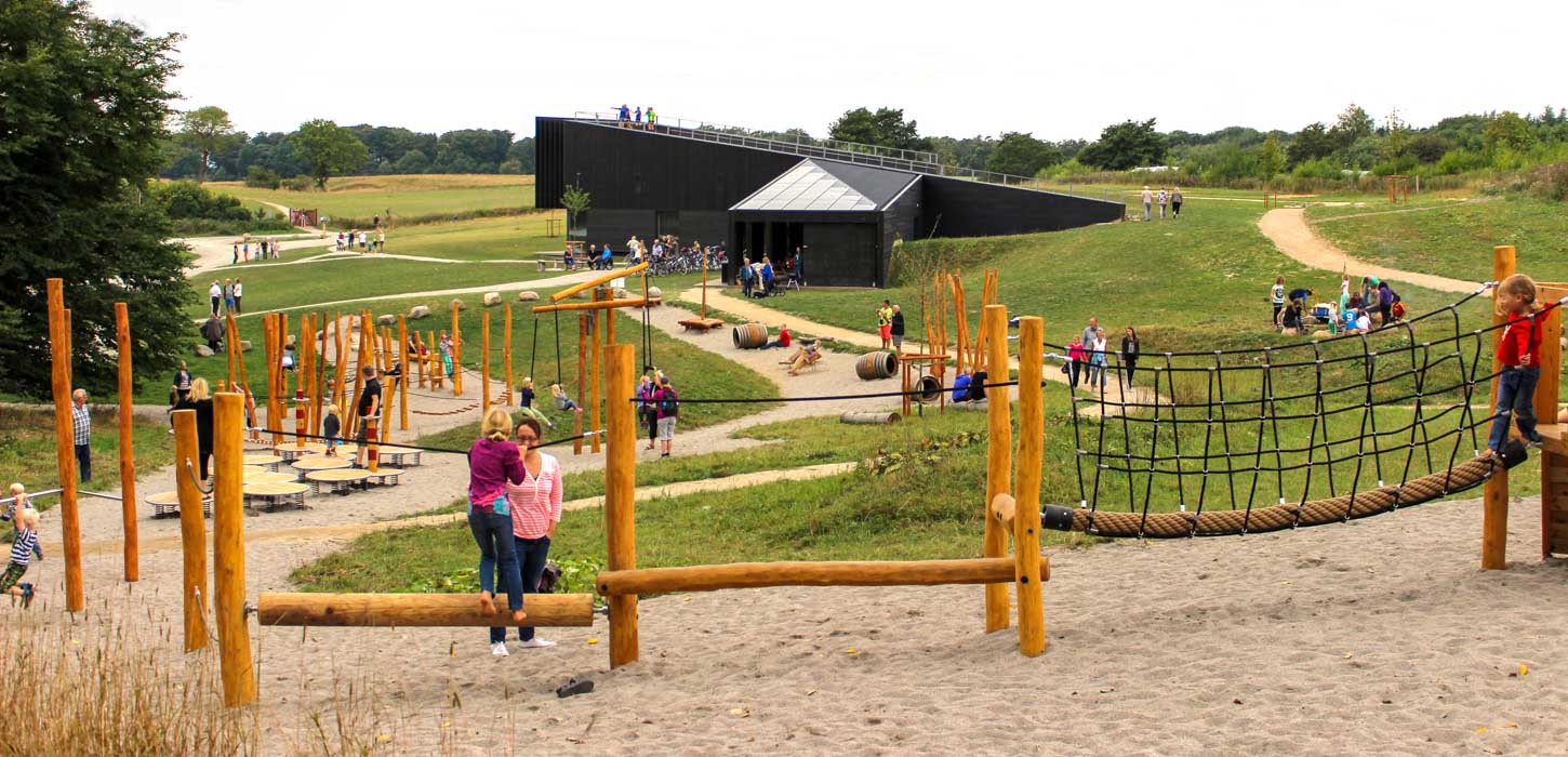 En lang forhindringsbane af træ snor sig ned midt i billedet. På den er en masse legende børn. Der er græs på begge sider og et stort sort hus i enden. 