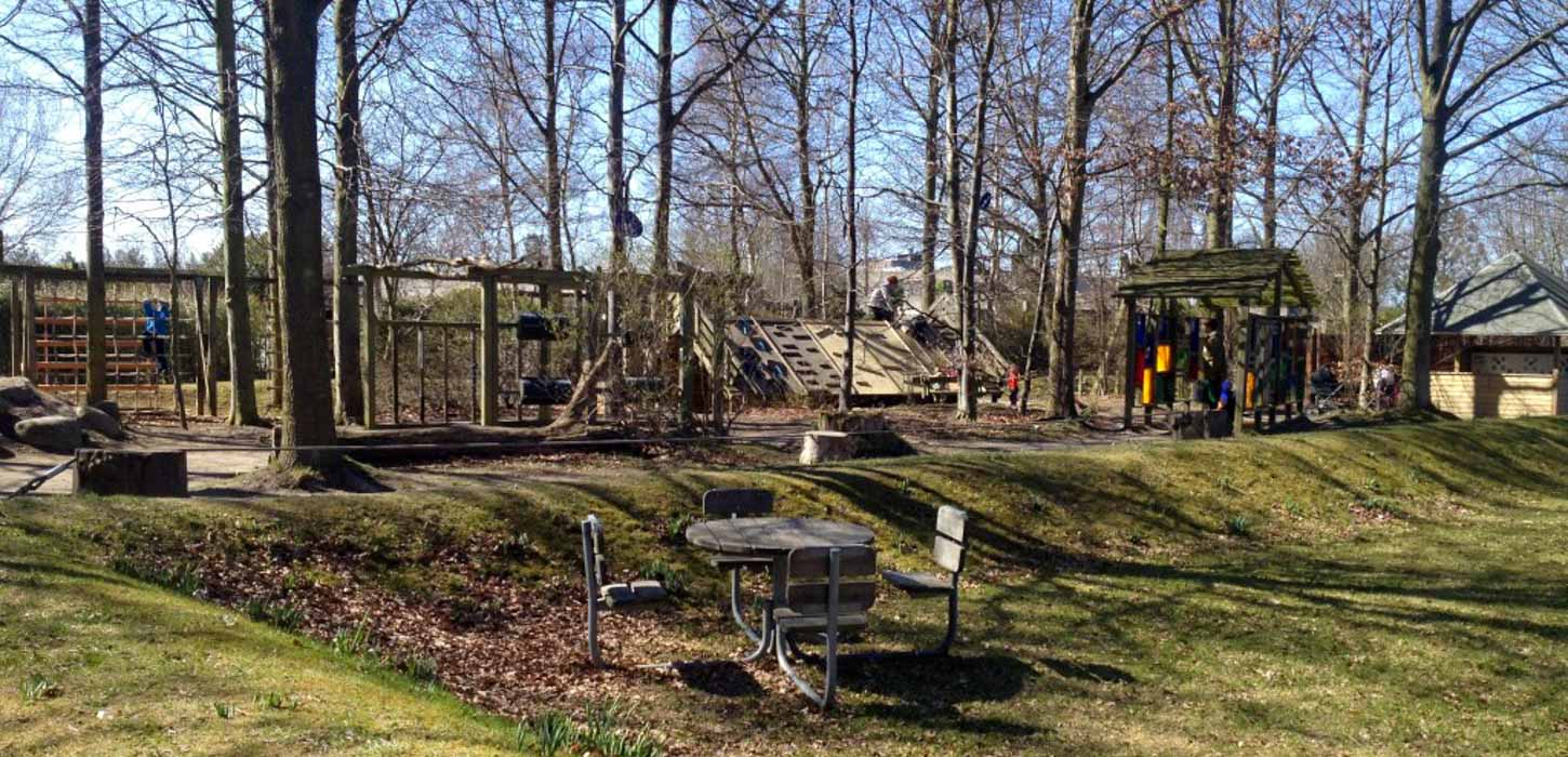 Ind mellem træerne i en skov ligger en stor legeplads af træ. Foran står en borde-bænkesæt.
