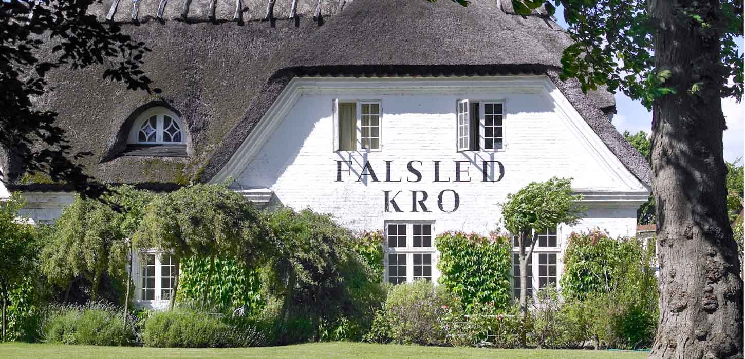 Hvid bygning med stråtag og en flot grøn have foran. Der står Falsled Kro på bygningen.
