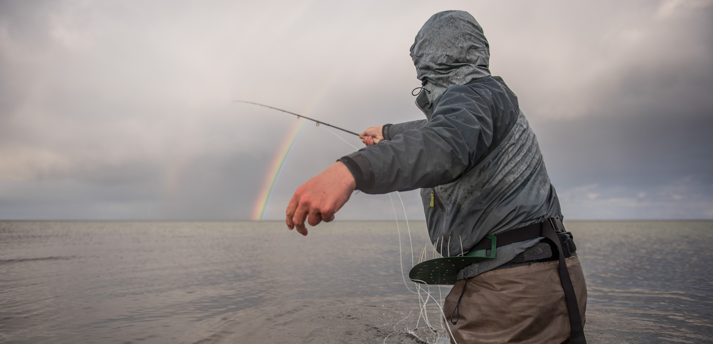 Lystfisker står med ryggen til og kaster ud. Han er iklædt mørkt tøj, og hætten på hans hoved er snørret godt ind. I baggrunden ses det sortgrå hav og en mørk uvejrshimmel, som en regnbue bryder igennem på.