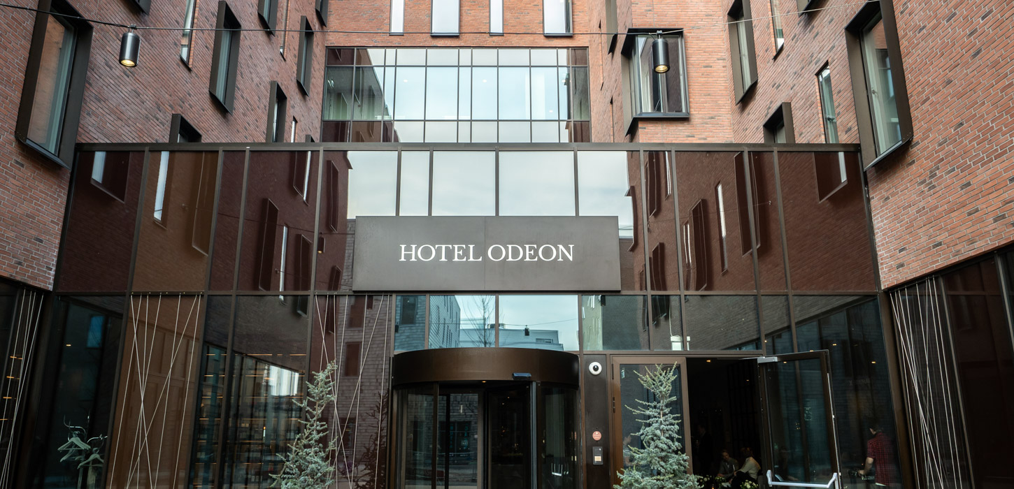 Ingangen til Hotel Odeon i Odense. En kobberfarvet svingdør er omringet af store vinduer. Over den hænger et skilt med hotellets logo.