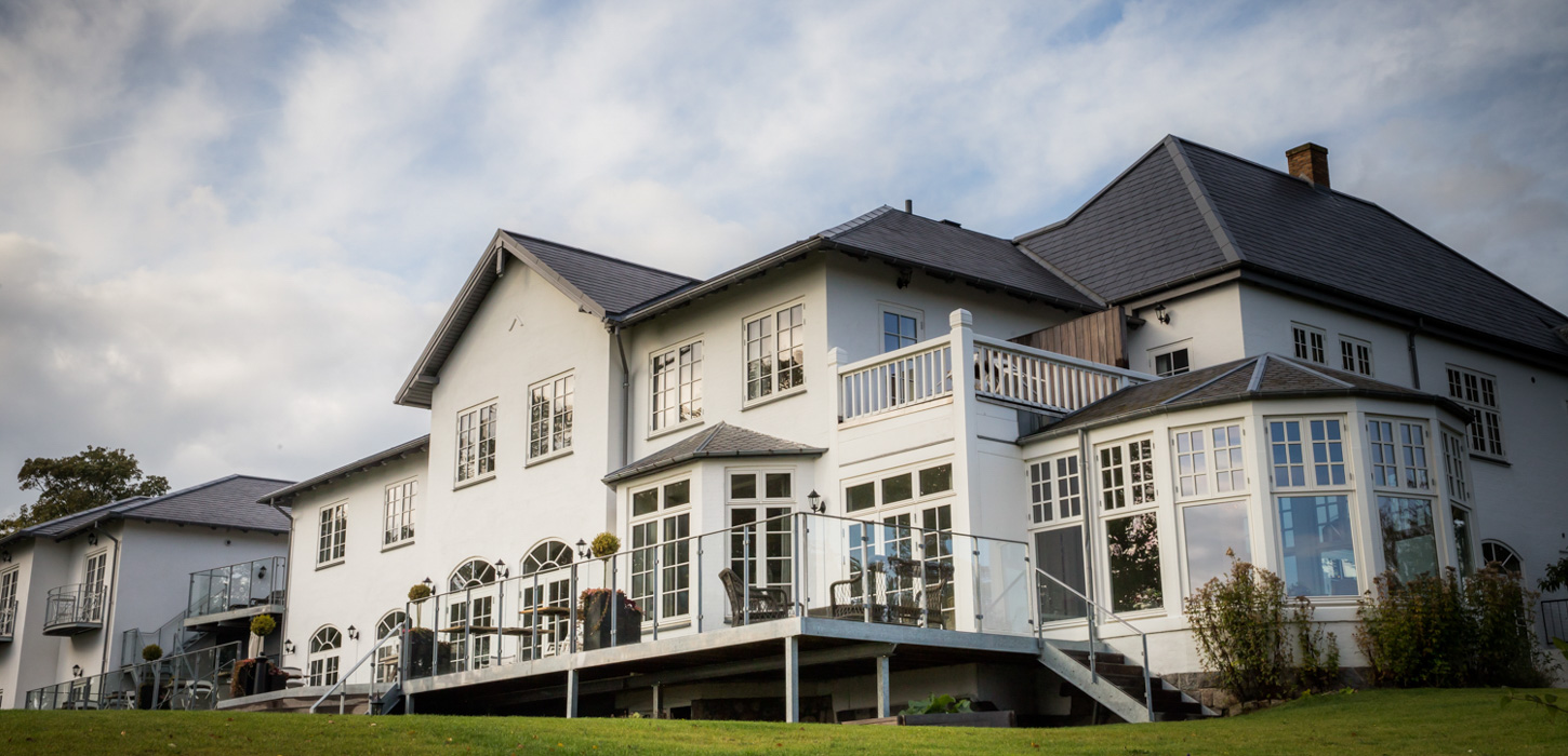 En billede taget nedefra af Hotel Stella Maris. Bygningen er hvid med mange vinduer og sort tag. Fra bygningen ligger en terrasse med sorte terrasse møbler. 