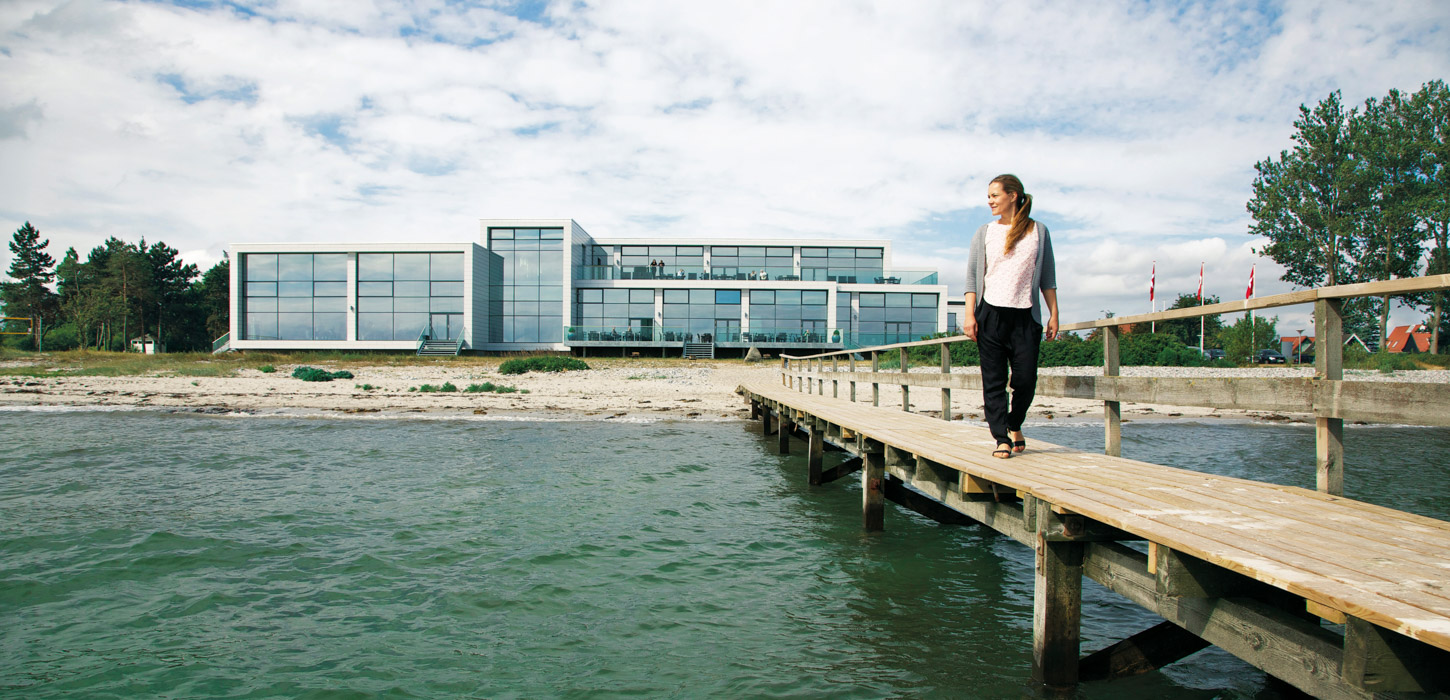 En dame går langs en badebro og kigger ud over vandet. Et stort hotel med vinduer på hele facaden ligger i baggrunden.