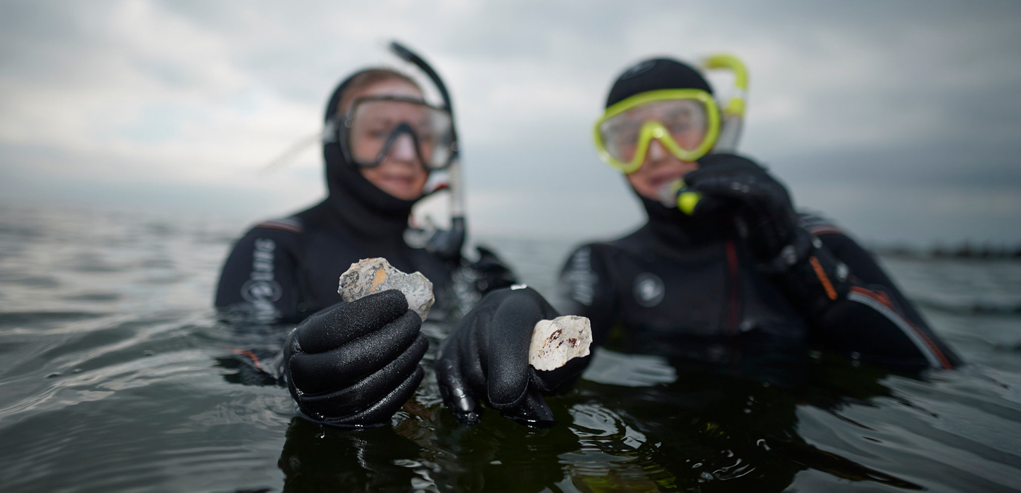 To personer i havet med snorkeludstyr viser fund fra havbunden frem. Der er overskyet og gråt.