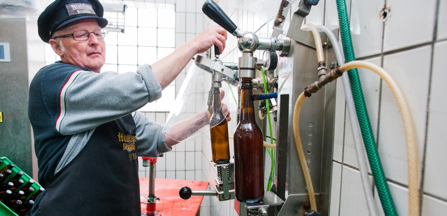 En brygger med høj hat står og fylder en ølflaske fra en taphane. Bag ham er bryggeriets hvide kakler og en ølkasse.