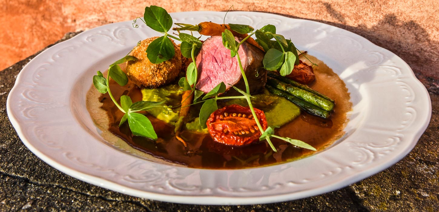 Nærbillede af smuk hvid tallerken med lækker anretning bestående af bl.a. et stykke rosa kød, bagt tomat, gulerødder, asparges og ærtespirer