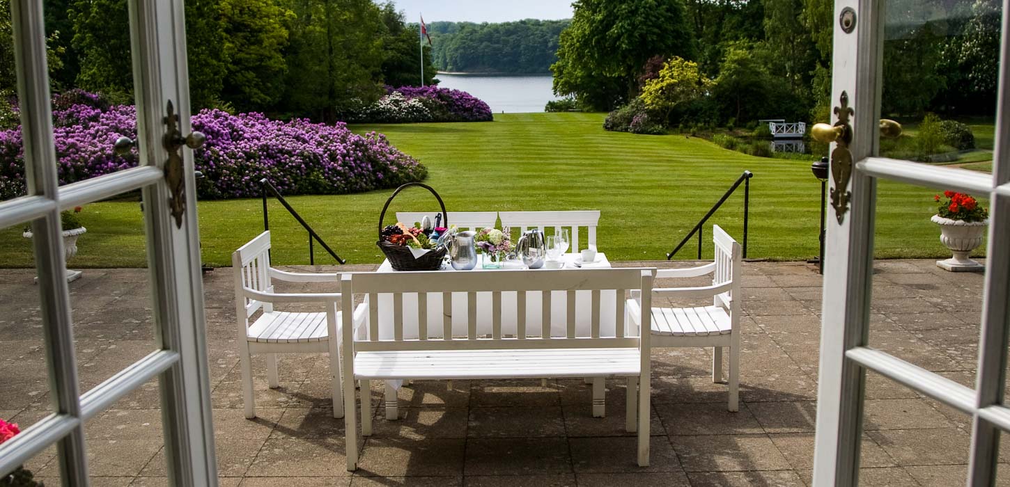 Hindsgavl Slots terrasse med hvide havemøbler. I baggrunden er en lang græsplæne med lilla blomsterbede der fører ned til vandet. 