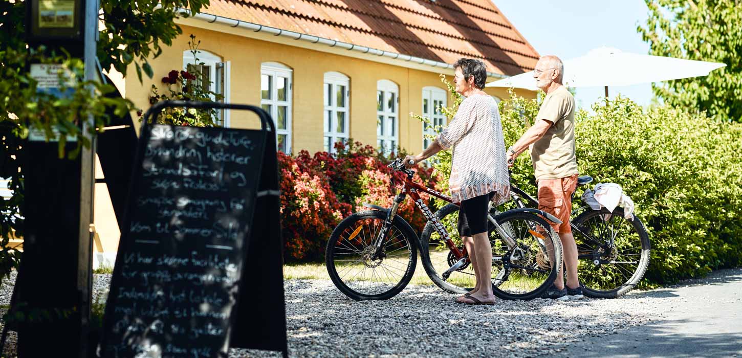 Mand og kvinde står afventende med deres cykler ved gult hus. 