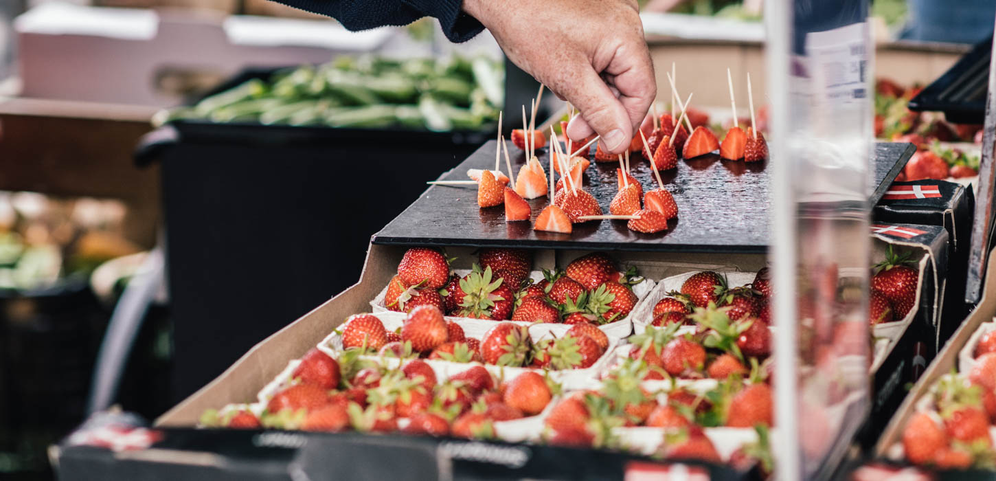 En sort kasse er fyldt med otte bakker jordbær. Oven på kassen står en sort bakke med udskårne jordbær med tandstikker i. En hånd er ved at tage et af disse jordbær. I baggrunden ses flere sorte kasser med ærter.