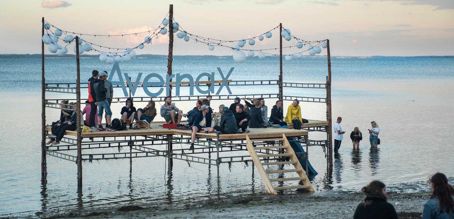 Åben Platform ude i vandet med lyskæder i toppen og Avanax-bogstaverne bagtil. Platformen trægulv er fyldt med siddende mennesker. Der står også tre mennesker ude i vandet.