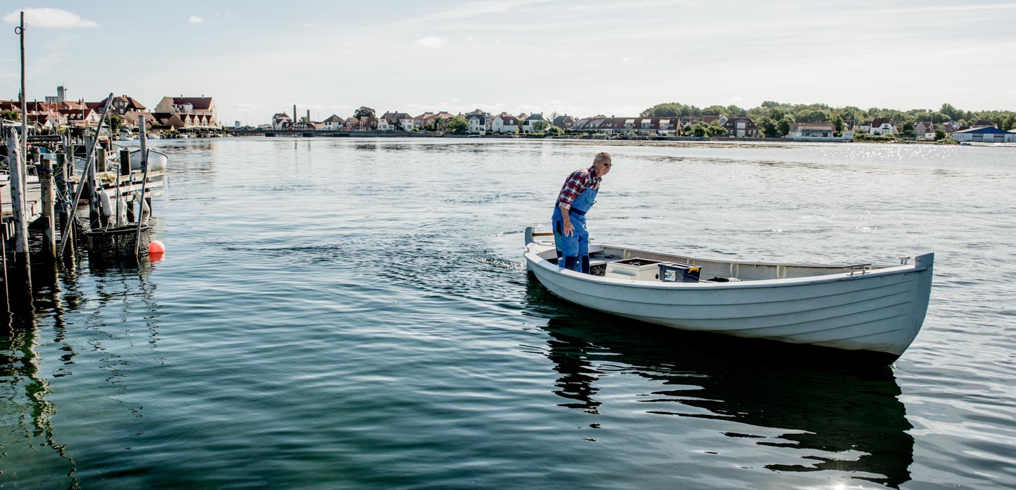 Hvid båd med mand i ternet skjorte ombord sejler ud fra Kerteminde Havn på roligt vand. Byen ses i baggrunden i horisonten.