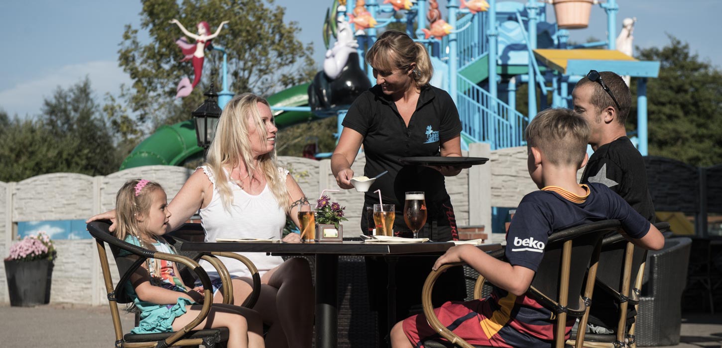En familie sidder ved et bord, hvor de får drikkevarer serveret. I baggrunden ses vandlandet på Bøsøre strand feriepark
