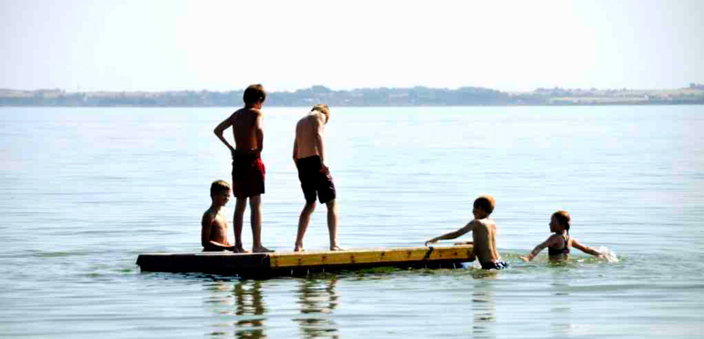 Fem badende børn ved Drejø. To børn står oppe på en træ-platform, de tre andre er nede i vandet.