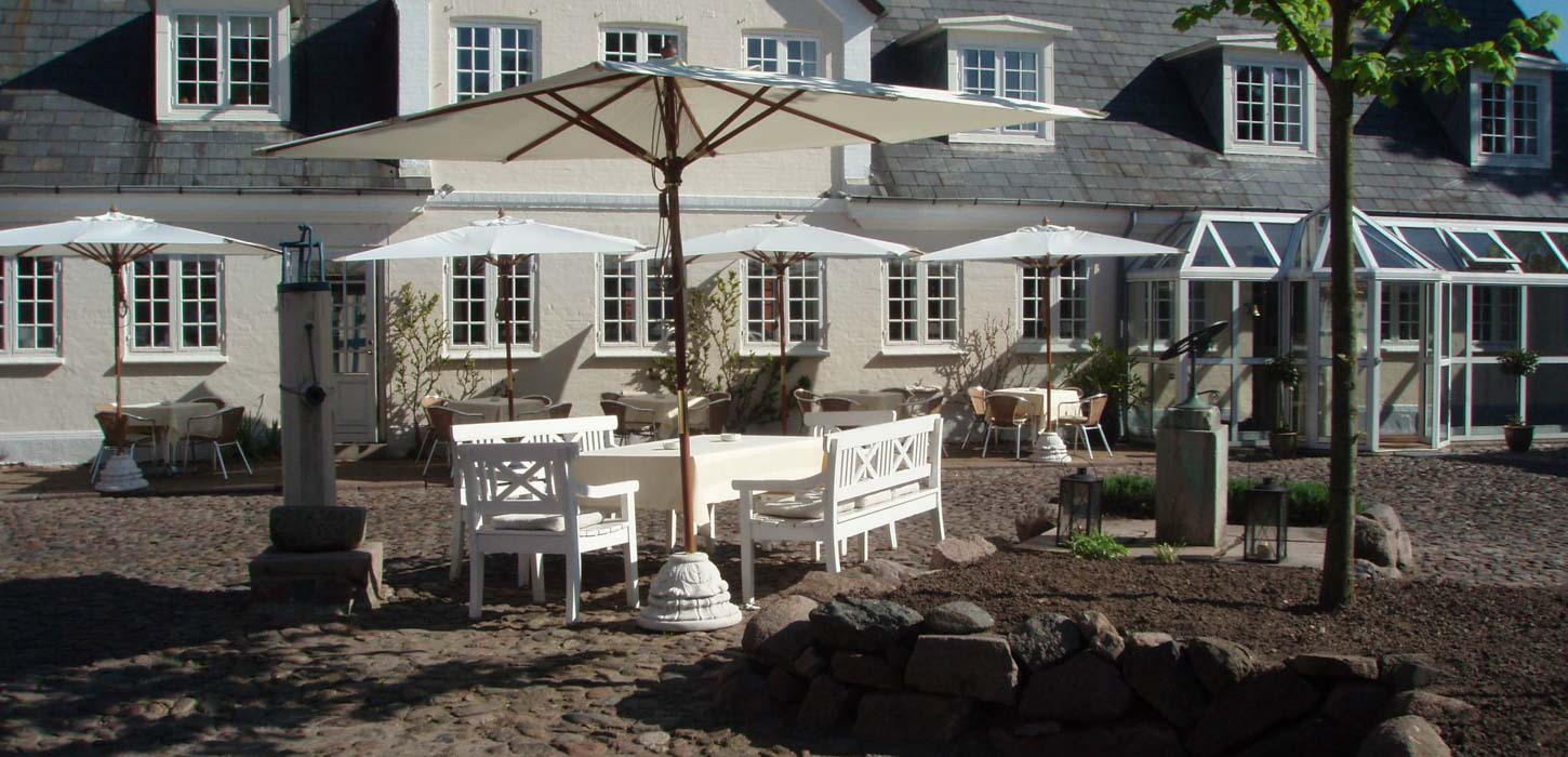 En stor terrasse på en solskinsdag med hvide borde, bænke og parasoller. Bagved er hotel og restaurant Knudsens Gård.