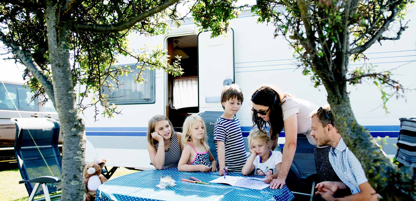 To voksne og fire børn samlet ved et campingbord foran en campingvogn