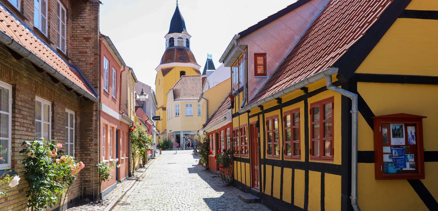 En malerisk gade i Faaborg med brostensbelægning og farverige huse med bindingsværk. Husene har levende gule og røde facader med traditionelle røde tegltage. I baggrunden tårner Faaborgs karakteristiske klokketårn sig op med sin røde kuppel og spir, hvilket tilføjer et charmerende historisk element til scenen. Det er en solrig dag med blå himmel og stokroser i fuldt flor, der tilføjer liv til gadebilledet.