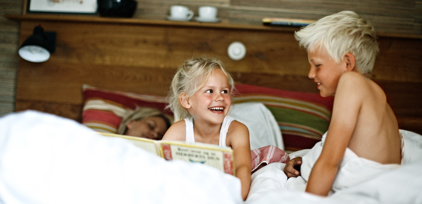 En lyshåret pige og dreng sidder i en seng, kigger på hinanden og griner. Pigen sidder med en bog i hånden.