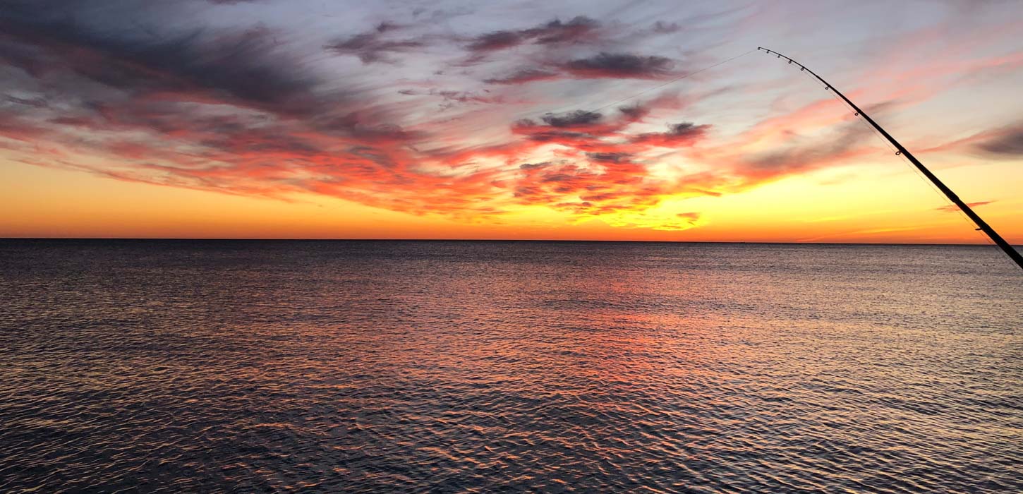 Den smukkeste gul-orange solnedgang ud over havet. I forgrunden til højre i billedet ses en fiskestang.