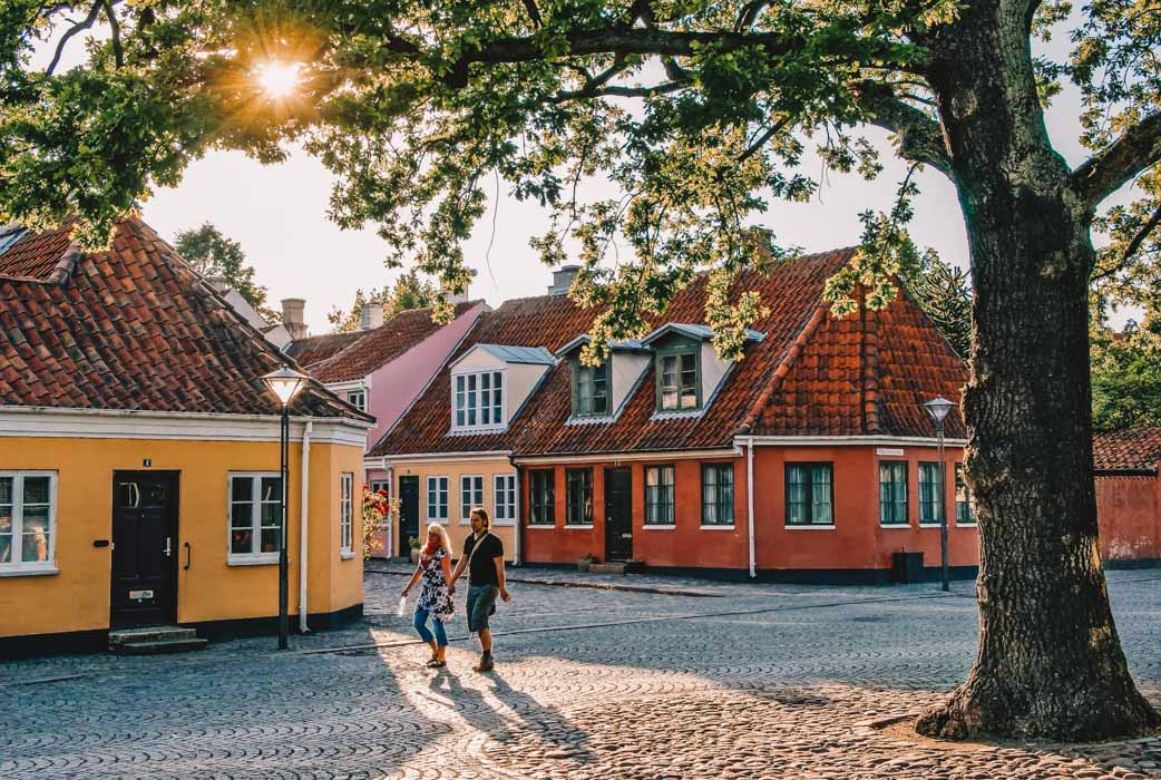 Et par går hånd i hånd på en brostensgade. I baggrundenden er fire bygninger i forskellige farver. Solen skinner gennem en trækrone. 