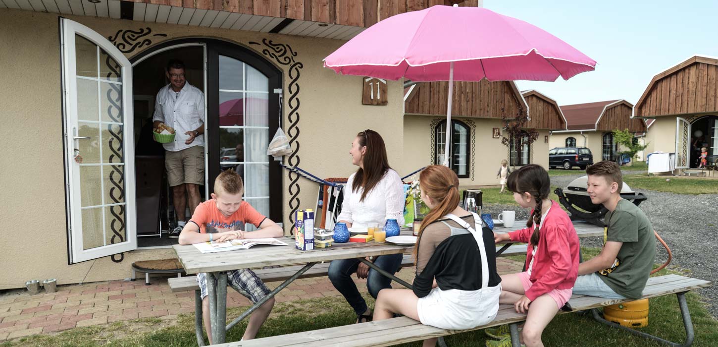 Ved et borde-bænke sæt sidder en familie og spiser aftensmad under en pink parasol. Ud fra sommerhuset i baggrunden kommer en mand med en skål i hånden. 