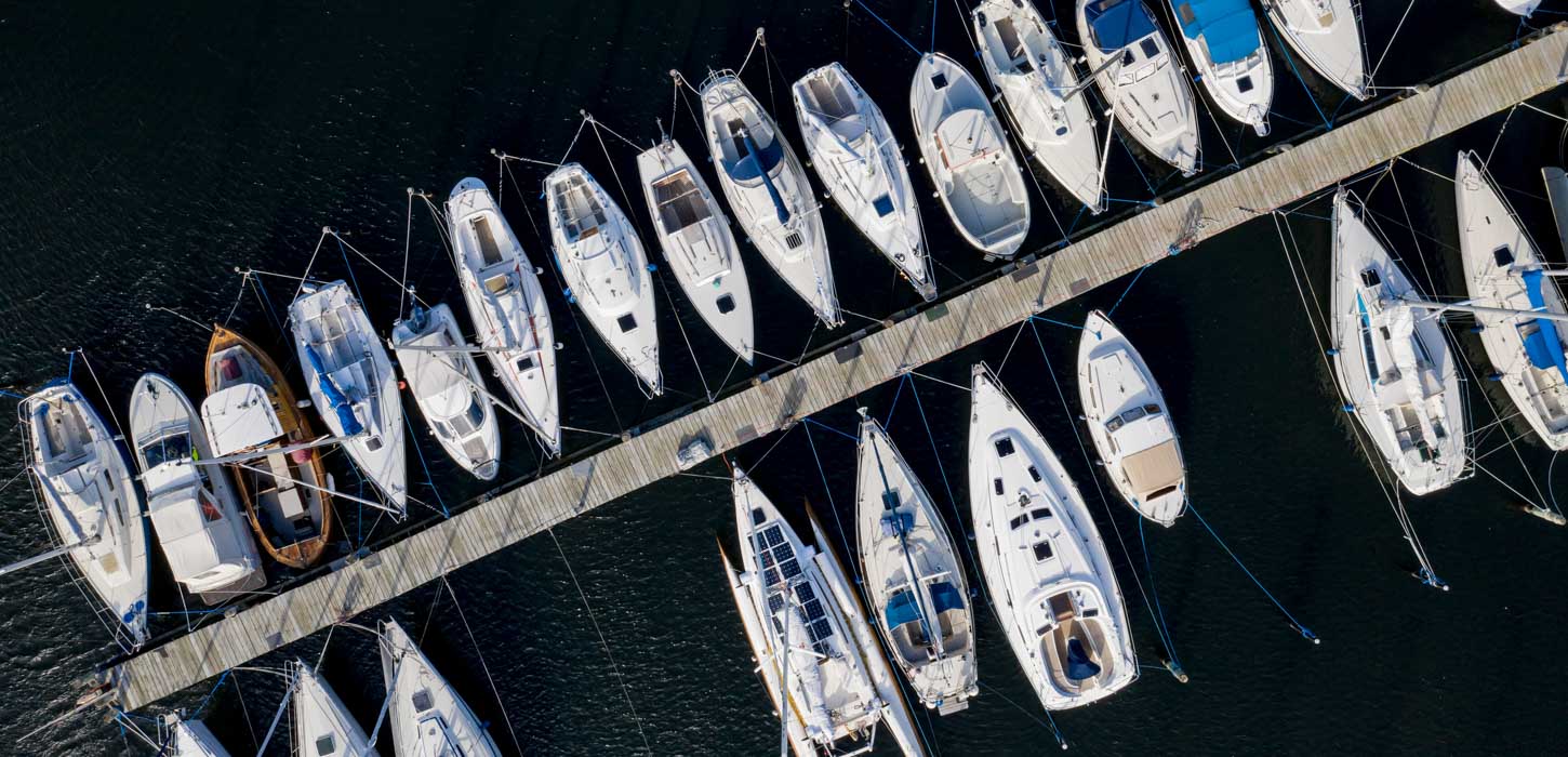 En lang havnebro ses oppefra. På begge sider er der fyldt med hvide både der ligger til kaj. Vandet omkring dem er mørkt.