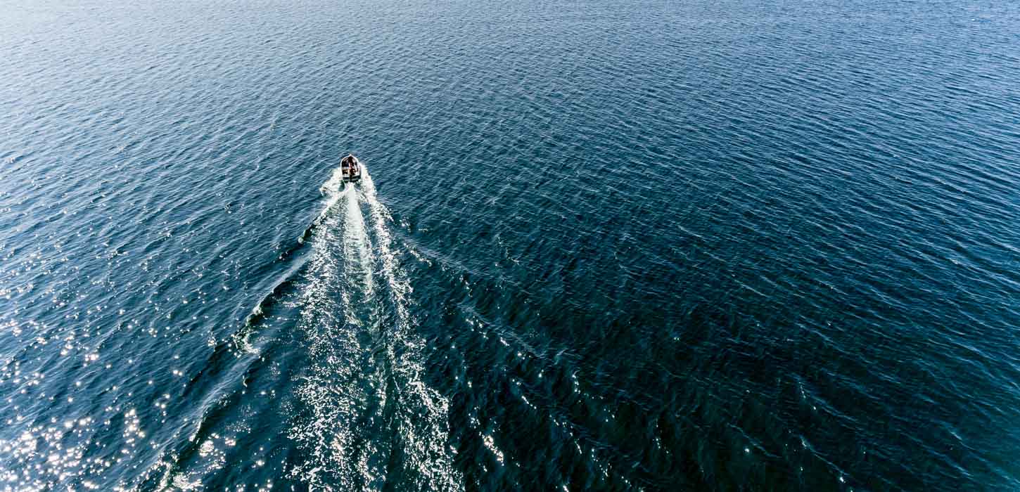 Dronebillede af en jolle motor båd sejler midt i det åbner hav. Bag den ses et sejlspor af hvidt havskum.