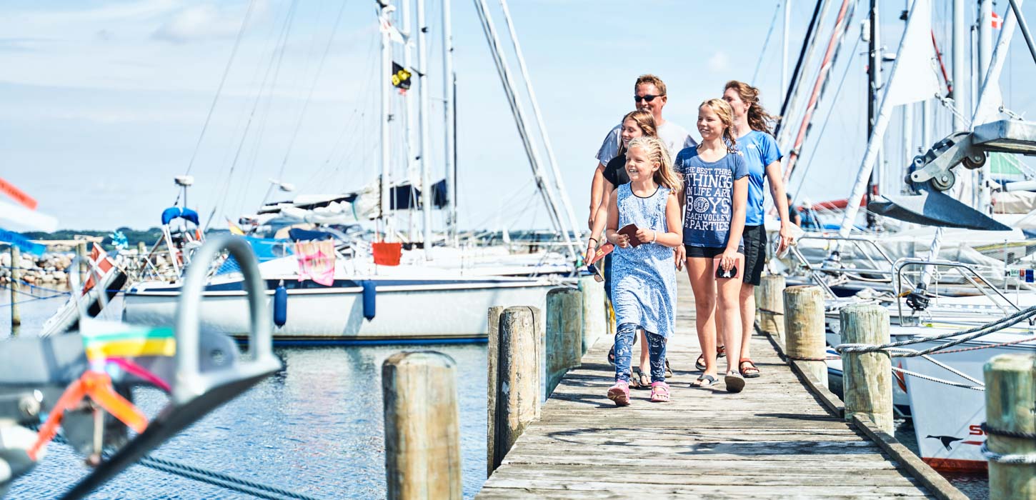 En familie kommer gående på en havn med en masse skibe i baggrunden.