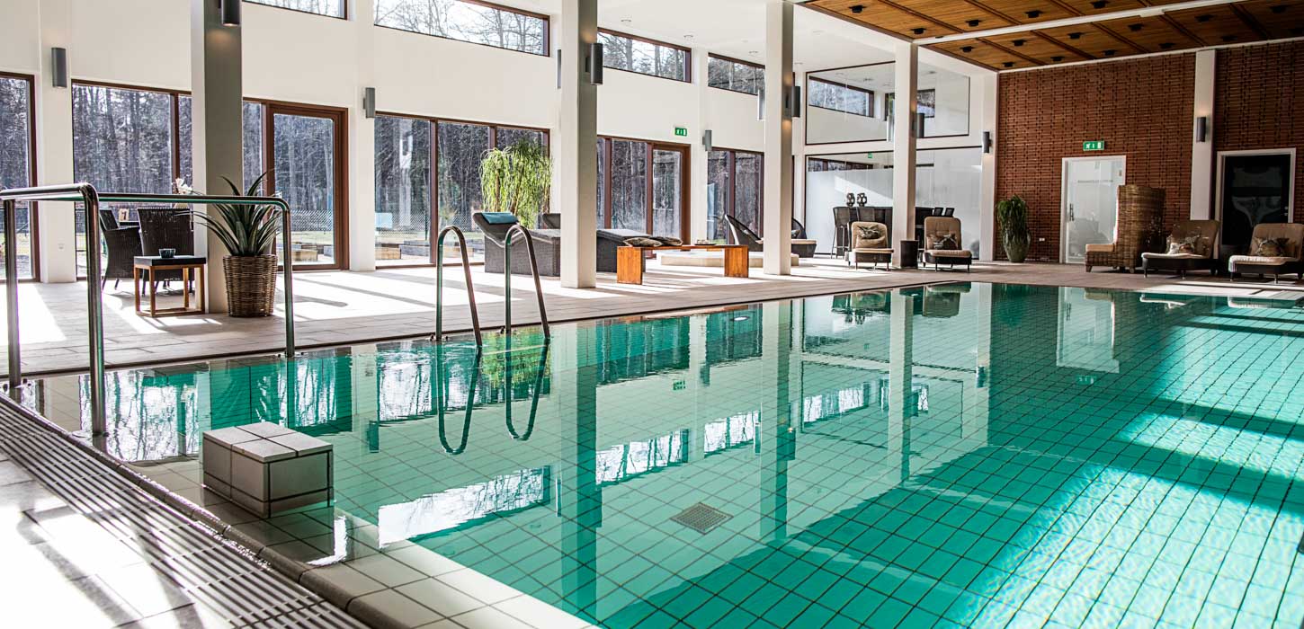 En swimmingpool med hvide klinker er omgivet af flettede liggestole. Der er store vinduer med udsigt til skov.  