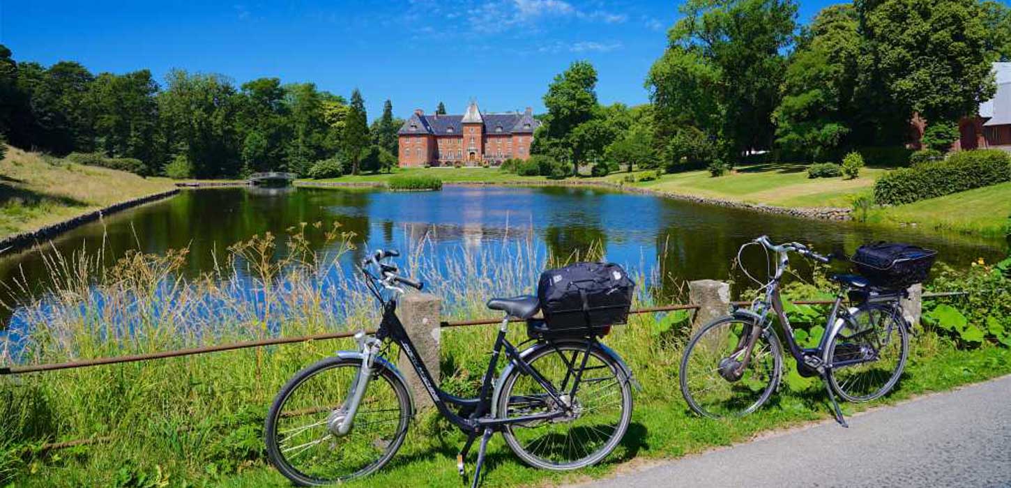 To cykler står parkeret ved noget vand. I baggrunden anes det røde Holstenhus.