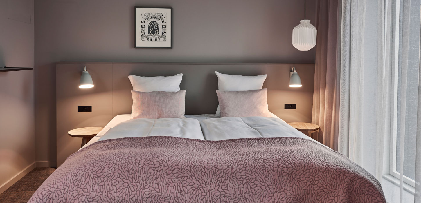 En dobbelt seng med lyserødt sengetæppe og pyntepuder. På hver side er der et sengebord af træ og en tændt læselampe. Over sengen hænger et billede på en grå væg.