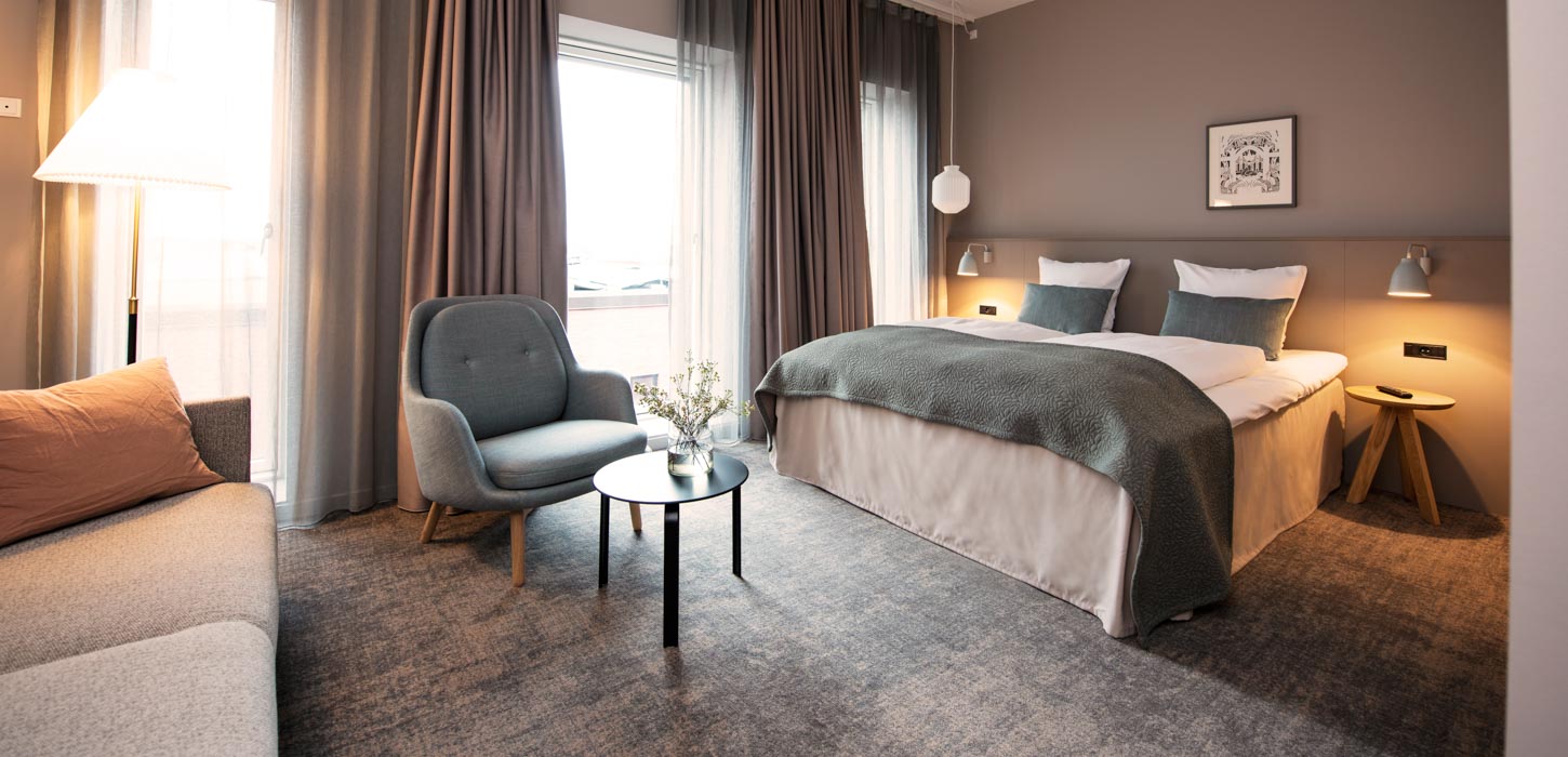 Et hotelværelse med brune vægge og et gråt gulvtæppe. Til venstre ser man hjørnet af en grå sofa, og en læne stol med et lille sofabord til. Til højre står en dobbelt seng med hvide lagner og blå pyntepuder. På hver side står der et natbord og en natlampe. 