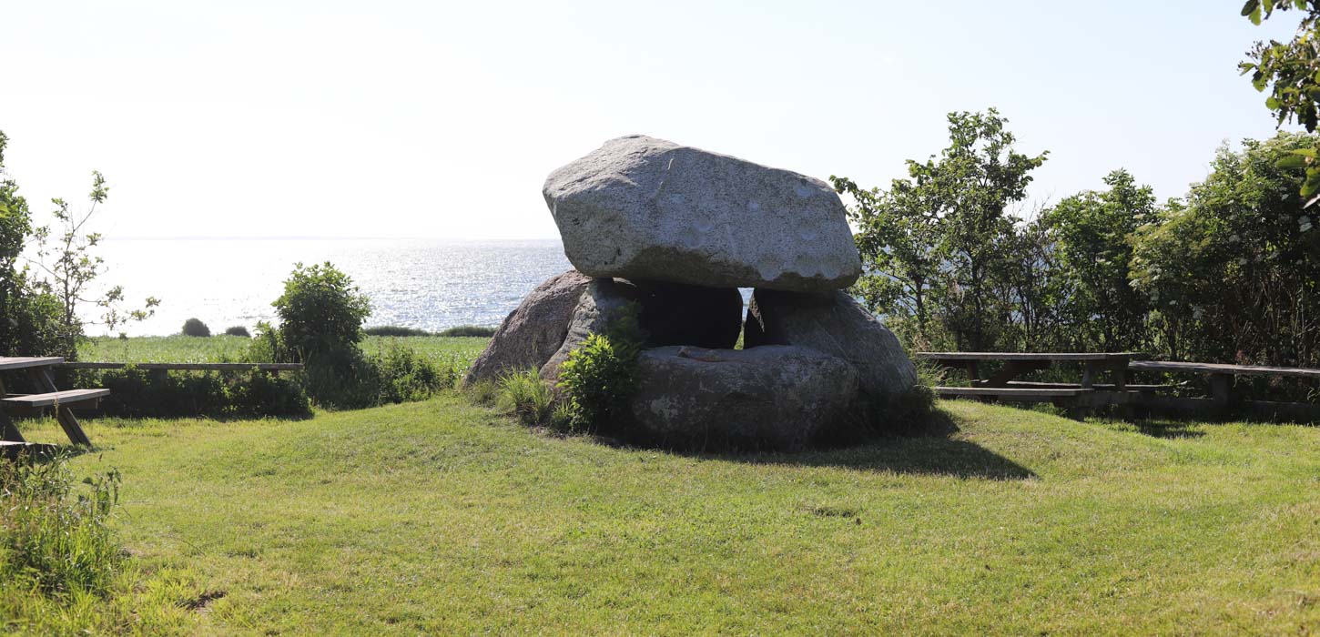 Billedet er af en græsplæne. I venstre side kan man se halvdelen af et bordbænke sæt, og i højre står et par træer. I midten af plænen er et dyssekammer af fire store sten. I horisonten kan man se havet.