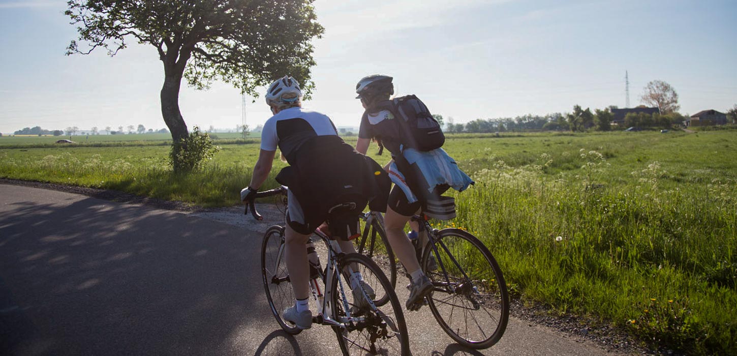 To cyklister i fart hen ad vejen. Træ og grønne marker i baggrunden.