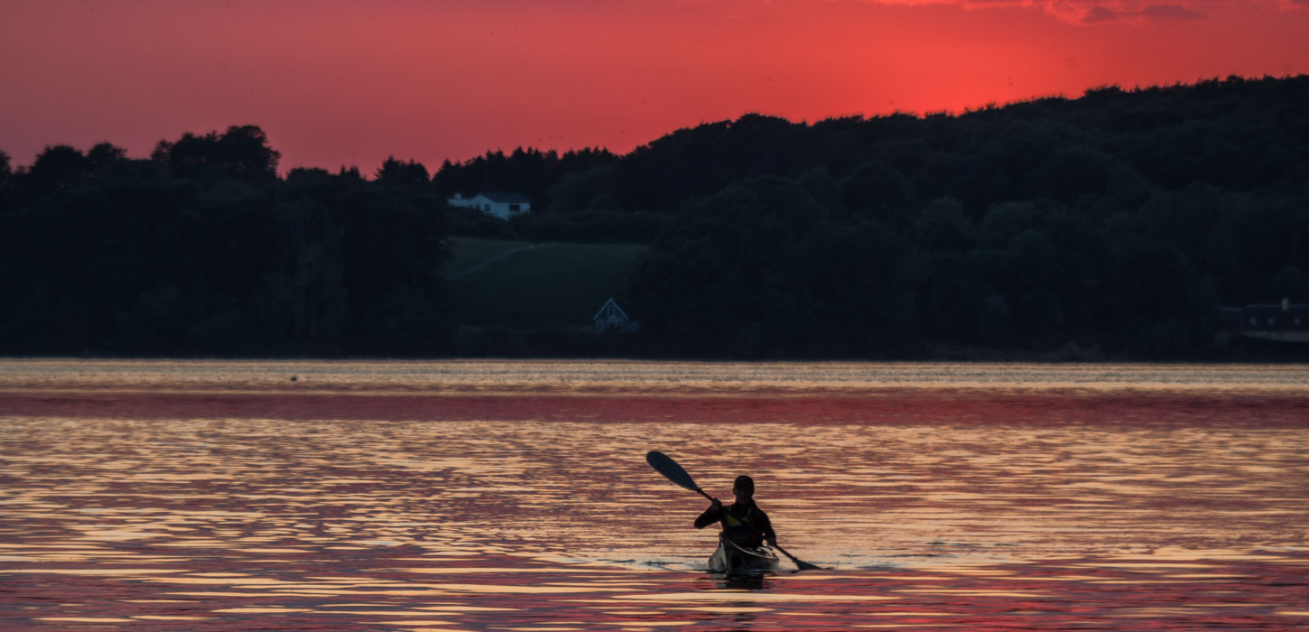 I baggrunden er himlen helt rød og foran er træer helt sorte da solen er på vej ned. Foran dette er der åbent hav hvor man kan se en enkelt man ro en kajak.