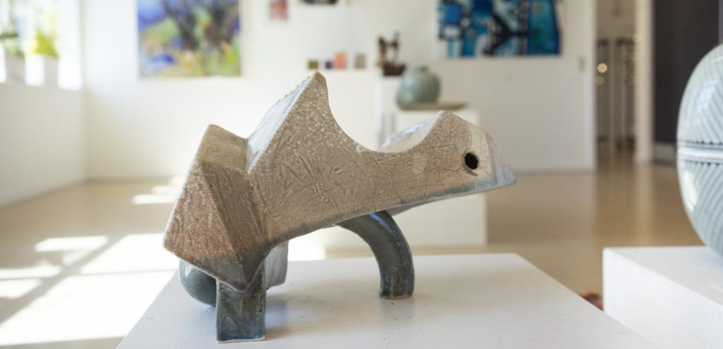 Et nærbillede af en kunstfigur på en hvid overflade. Figuren ligner en skildpadde, hvor man har gjort alle runde kanter skarpe. I baggrunden ses resten af et kunstmuseum. 