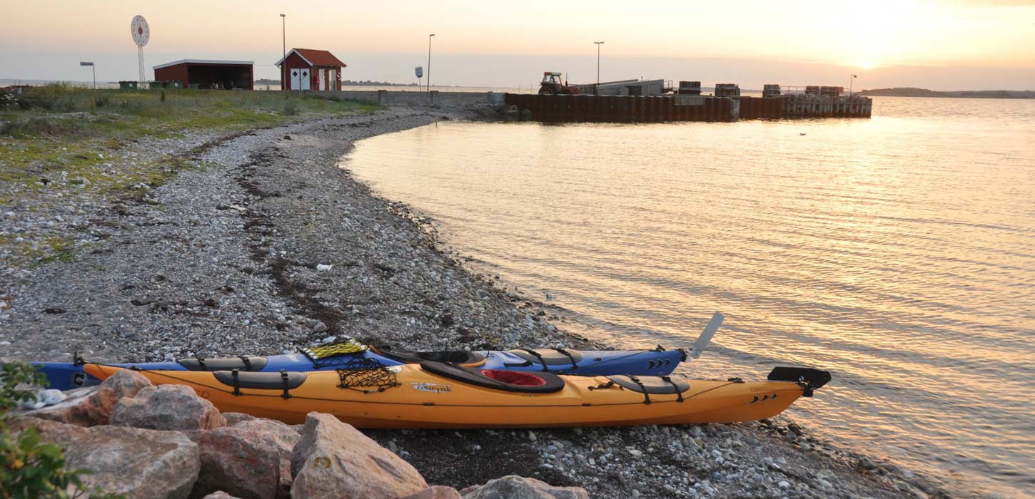 På en strandbred ligger to kajakker i forgrunden. I horisonten af billedet kan man se en  strandspids hvor der står et lille rødt hus foran solnedgangen. 