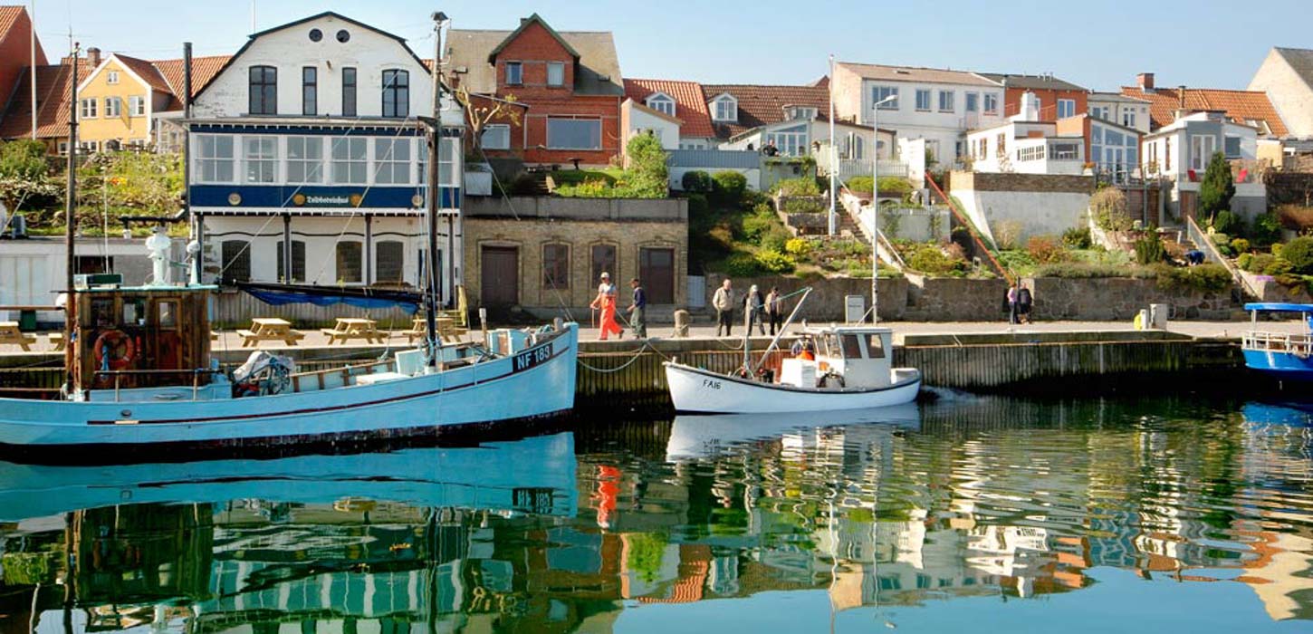En havn set fra vandet. I vandet er der en lille jolle og en stor gammel sejlbåd. På en bakke over havnen er der placeret gamle bygninger, der har udsigt til havnen.