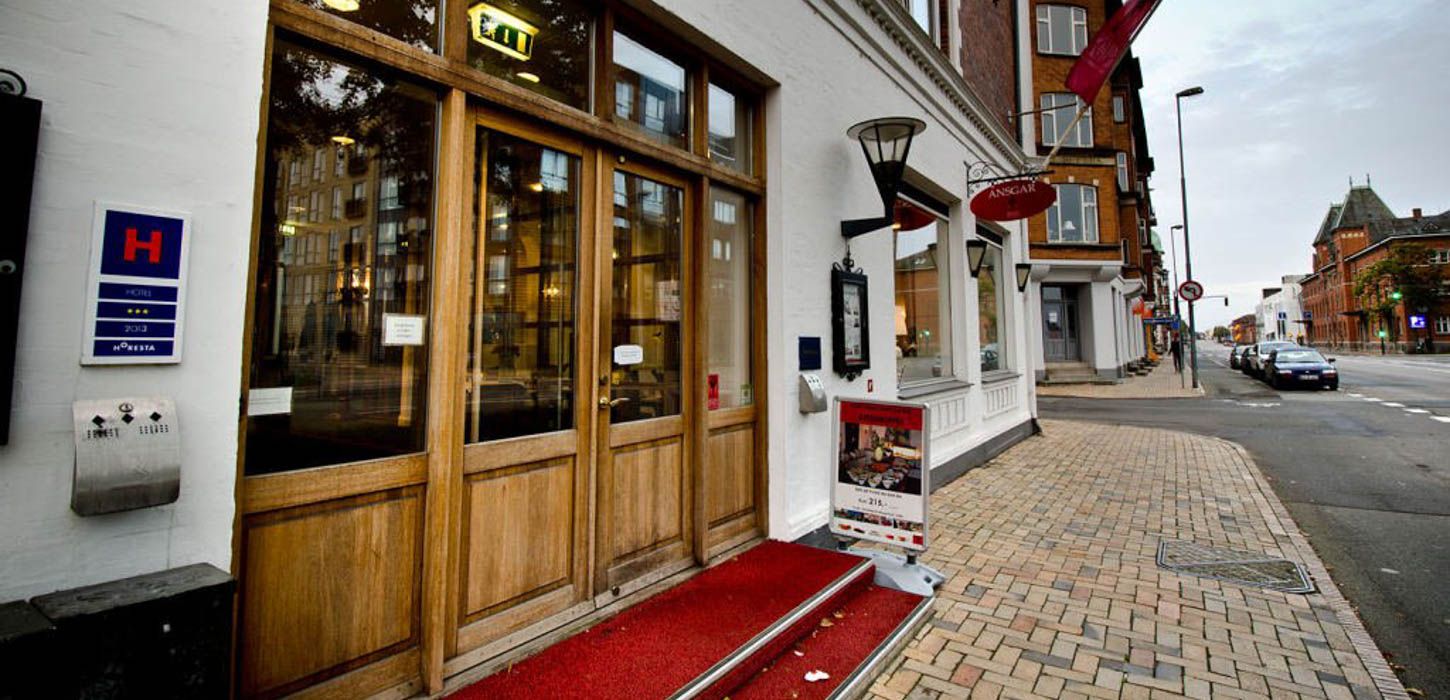 Indgangsparti af mørkt træ. Foran er en trappe med to trin og røde farver og så er der sat et skilt ud på gade som reklame for stedet