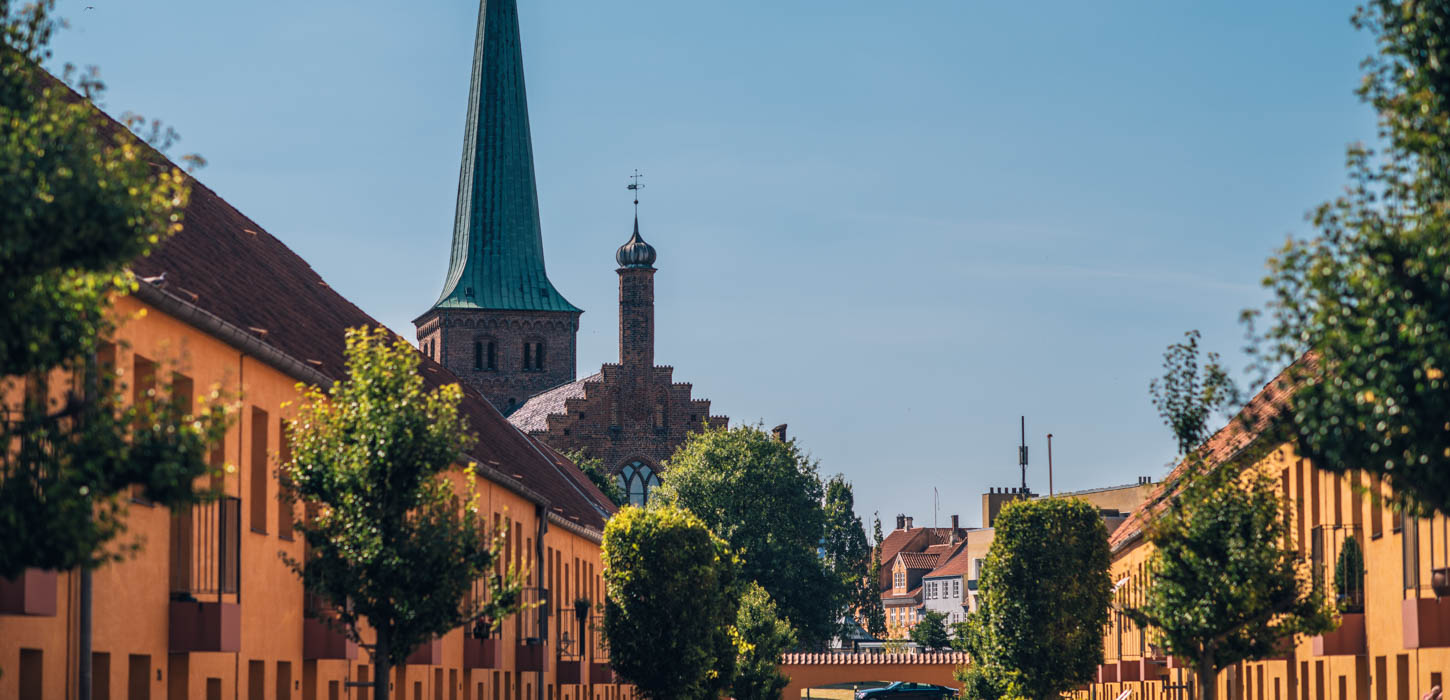 I forgrunden ses toppen af en lang række gule huse med et par træer foran. I baggrunden ses en blå himmel med et kirketårn foran.