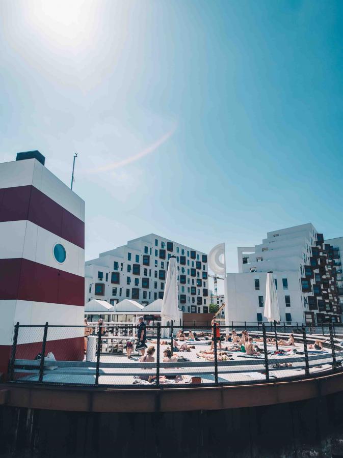 Odenses havnebad på en sommerdag. De besøgende i havnebadet ligger og soler sig i det gode vejr. I baggrunden er der hvide lejligheder.