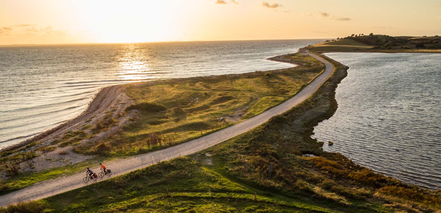 To cyklister set oppefra på vej henover grøn tange med vand til begge sider.