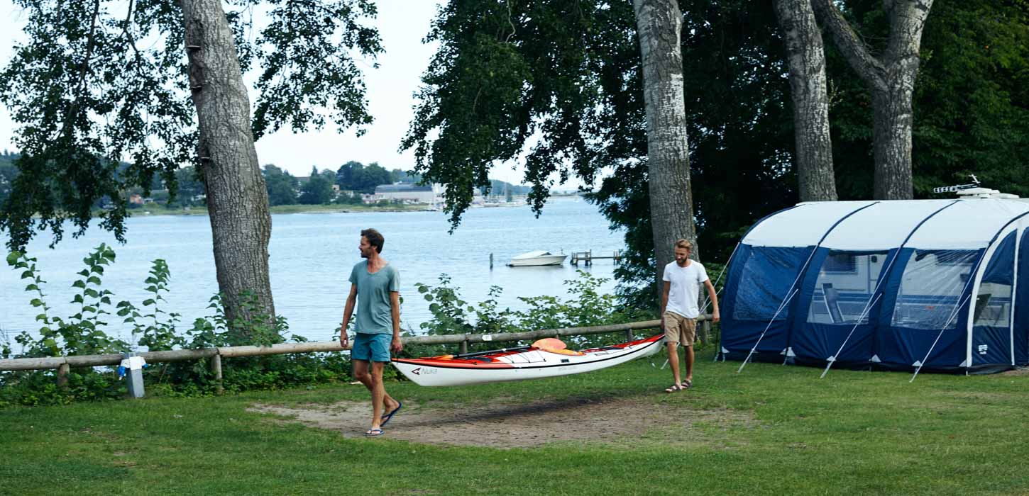 To mænd i bare arme og ben kommer gående, bærende på en kajak. Store træer og vand i baggrunden. Et telt ses i den ene side.