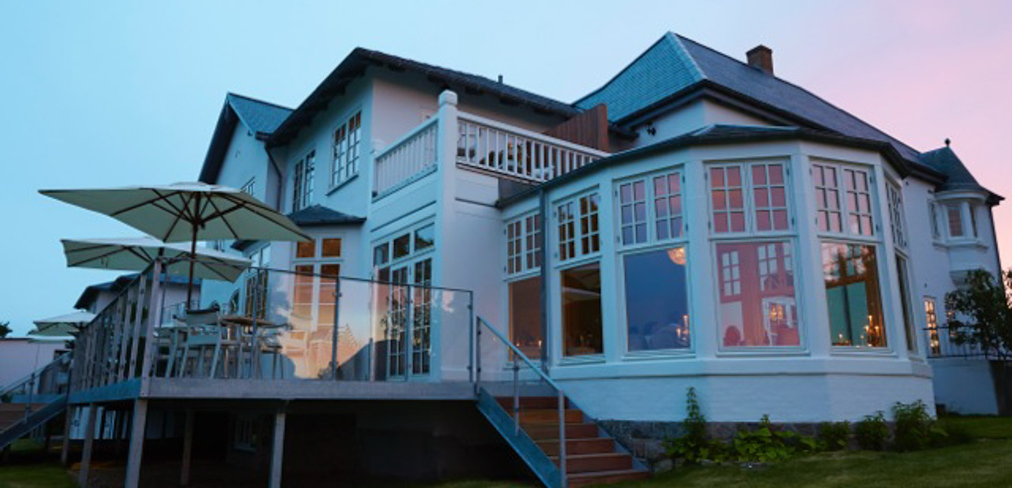 Hvidt hotel med store vinduer fotograferet nedefra med en flot lyseblå og lyserød himmel i baggrunden.