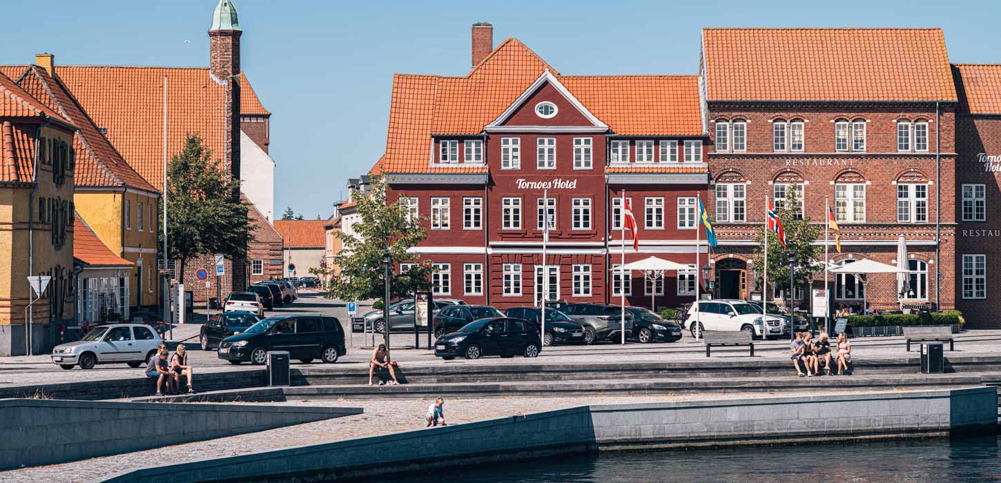 Store, røde huse foran havnefronten i Kerteminde. I forgrunden sidder der folk og nyder solen, en masse parkerede biler i baggrunden.