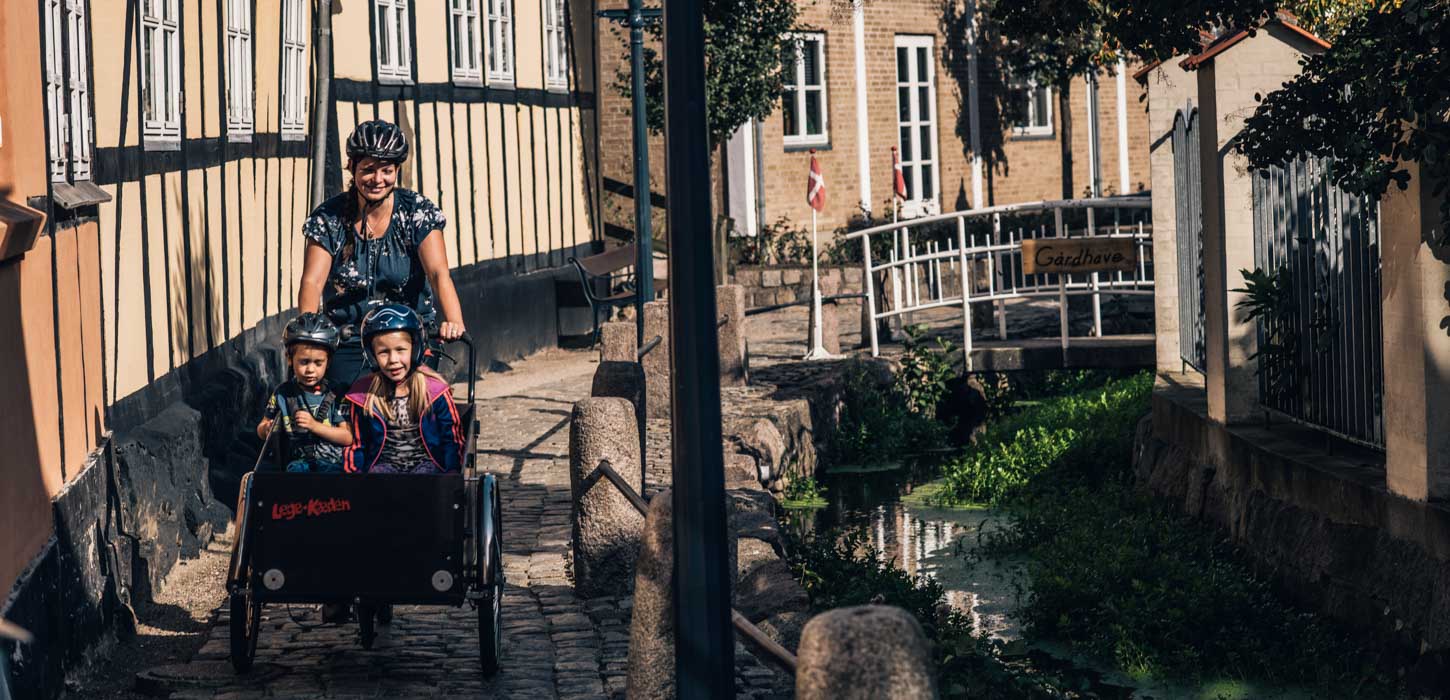 Kvinde kommer kørende på ladcykel med to børn i på en smal, brostensbelagt gade med Bybækken til den ene side og gamle bindingsværkshuse til den anden side.