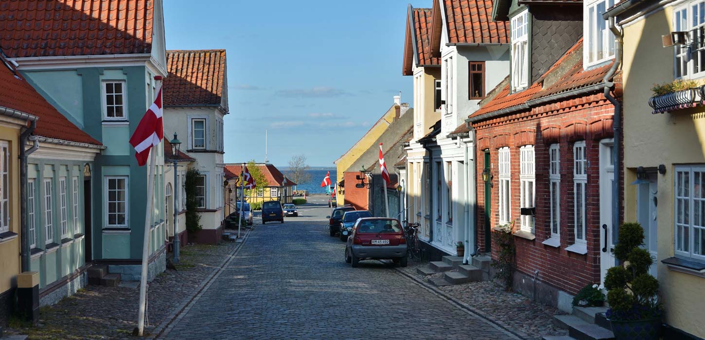 Brostensbelagt gade med gamle byhuse i forskellige farver til begge sider. Der er flag sat op foran et par af husene, et par parkerede biler, og den blå himmel ses i forsvindingspunktet.