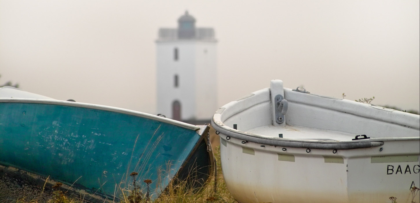 I forgrunden ses to både, der ligger og er trukket op på land, en havblå og en hvid. I en sløret baggrund ses Bågø Fyr.