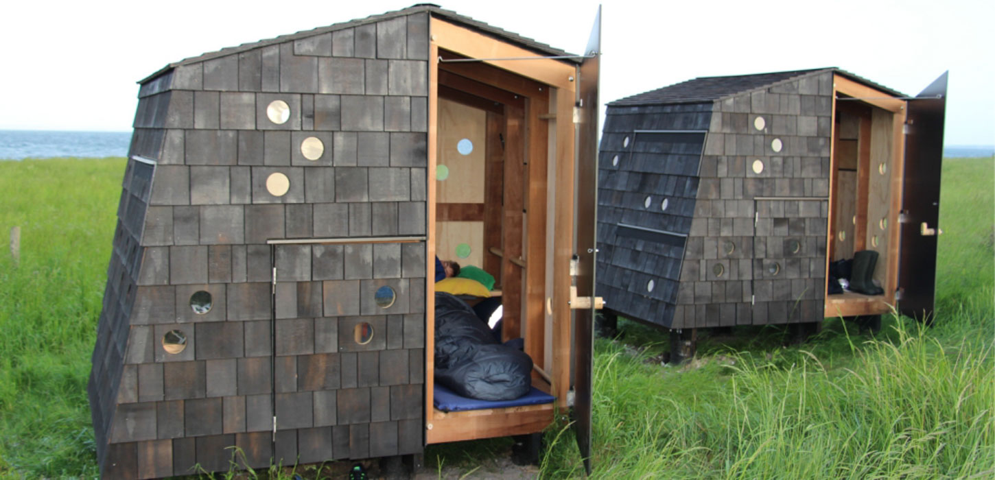 To shelters midt i en grøn eng med svag havudsigt i baggrunden.